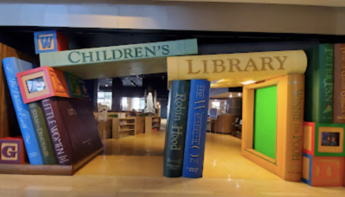 Colorful oversized books form an archway labeled "Children's Library," leading into a cozy reading space.