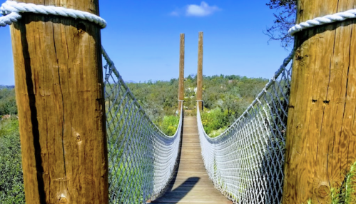 A wooden suspension bridge with rope railings stretches over a scenic landscape under a clear blue sky.
