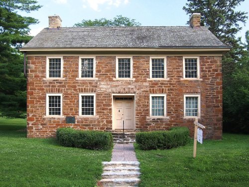 A historic stone house with a symmetrical facade, surrounded by grass and trees, featuring a central door and windows.
