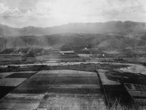 Aerial view of a river winding through fields and hills, with a mix of farmland and natural landscape in black and white.