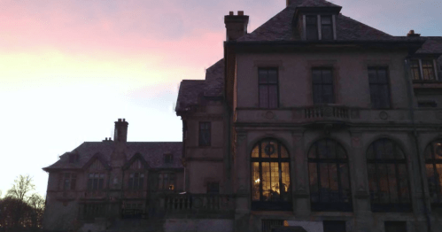 A large, historic mansion at sunset, with intricate architecture and warm light glowing from the windows.
