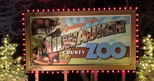 A colorful sign reading "Greetings From Milwaukee County Zoo" with bear illustrations, framed by festive lights.