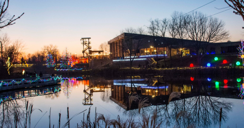 A serene evening scene with colorful lights reflecting on a calm pond, surrounded by trees and a building.