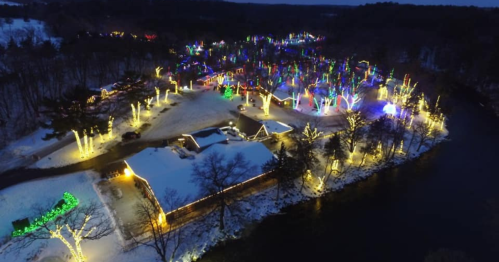 Aerial view of a snowy landscape adorned with colorful holiday lights, illuminating trees and buildings along a river.