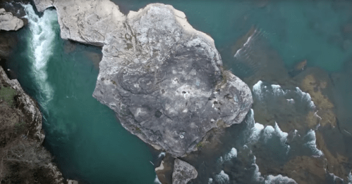 Aerial view of a large rock surrounded by flowing turquoise water and small rapids.