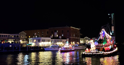 Colorful boats decorated with lights float on a dark river, with a festive waterfront building in the background.