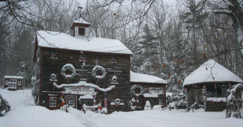 A snowy scene of a rustic wooden building decorated for the holidays, surrounded by trees and a winter landscape.