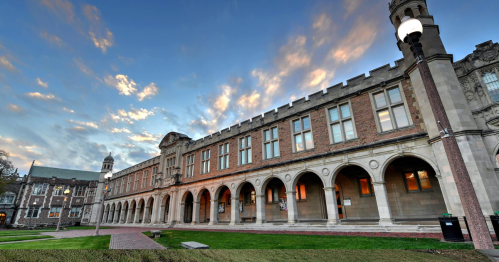 Historic building with arched windows and a stone facade, set against a colorful sunset sky. Lush green lawn in front.