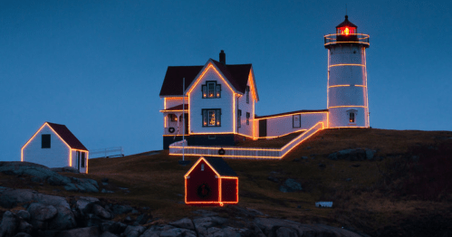 A lighthouse and house decorated with orange lights, set against a twilight sky. Rocks in the foreground.