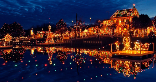 A beautifully lit house and trees adorned with colorful Christmas lights, reflecting in a calm water surface at dusk.