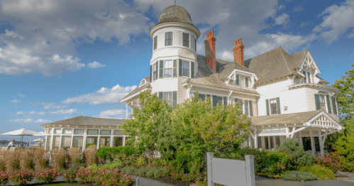 A large, elegant white mansion with a turret, surrounded by greenery and flowers, under a blue sky with clouds.