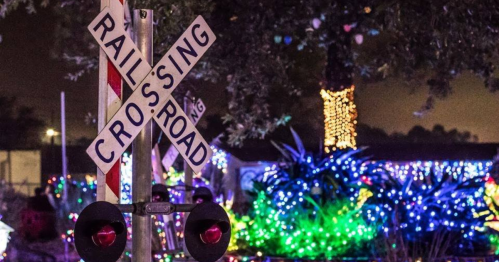 A railroad crossing sign in front of colorful holiday lights and decorations at night.