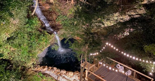 A serene outdoor scene featuring a waterfall, a pond, and a deck with string lights surrounded by lush greenery.