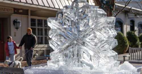 A large, intricate ice sculpture of a snowflake sits in a winter scene, with people walking a dog in the background.