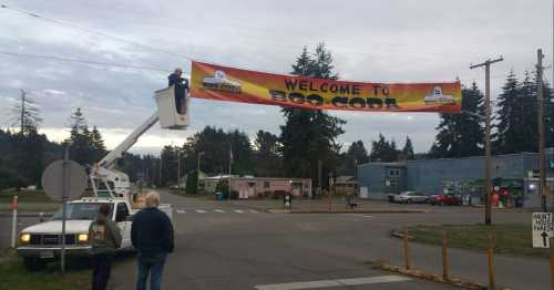 A worker in a lift hangs a "Welcome to Boo-Cora" banner above a street, with trees and buildings in the background.