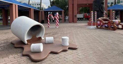 A large, tipped-over cup spilling hot chocolate with marshmallows on a brick plaza, surrounded by festive decorations.