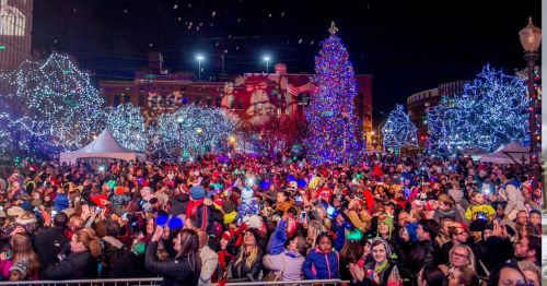 A large crowd gathers at night, celebrating around a brightly lit Christmas tree and festive decorations.