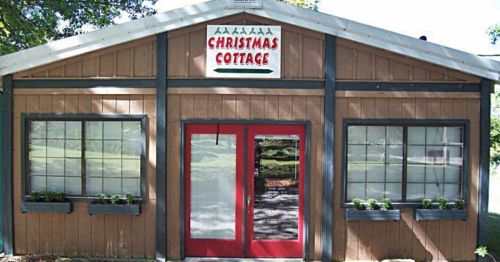 A wooden building with a sign reading "Christmas Cottage," featuring red doors and window boxes with plants.