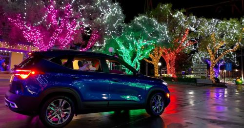 A blue car parked near colorful, illuminated trees with vibrant holiday lights in a festive outdoor setting.