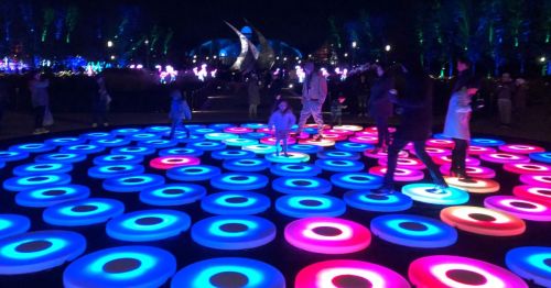 A vibrant outdoor scene with people walking on illuminated circular patterns in blue, pink, and white at night.