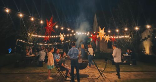 A lively outdoor gathering at night, with string lights and colorful stars, people enjoying a fire and each other's company.