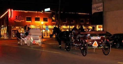 Two horse-drawn carriages decorated with lights travel down a street at night, with shops illuminated in the background.