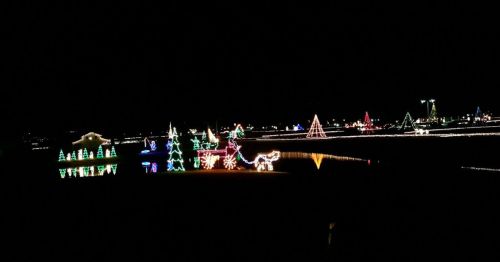 Colorful holiday lights illuminate a dark landscape, featuring trees, reindeer, and festive decorations reflected in water.