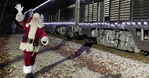 Santa Claus waves beside a decorated train on a gravel track, surrounded by trees and holiday lights.