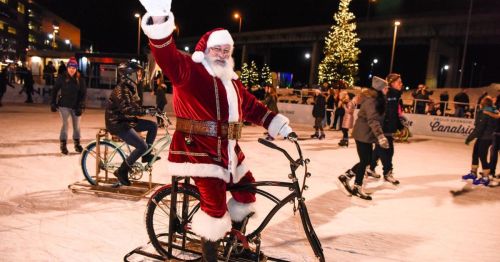 Santa Claus waves while riding a bike on an ice rink, surrounded by festive lights and people skating.