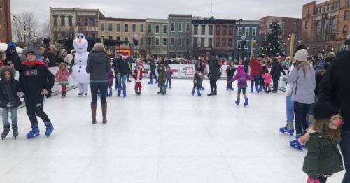 A busy outdoor ice skating rink with families and children skating, festive decorations, and a snowman character nearby.