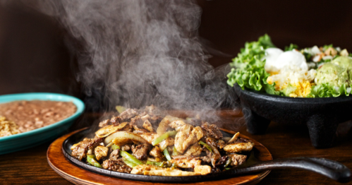 Sizzling fajitas with beef and peppers, steam rising, alongside a fresh salad and beans on a wooden table.