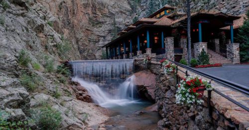 A scenic view of a building beside a waterfall, surrounded by rocky cliffs and lush greenery.