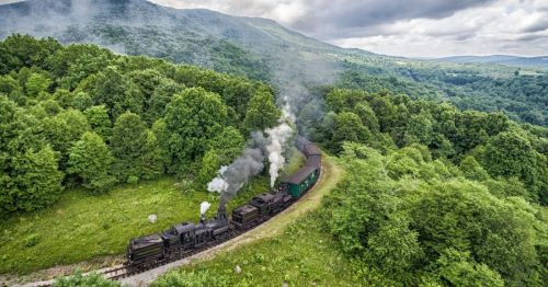 A vintage steam train winds through lush green hills under a cloudy sky.
