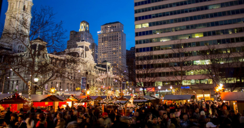 A bustling holiday market at dusk, with festive lights and crowds, surrounded by historic and modern buildings.