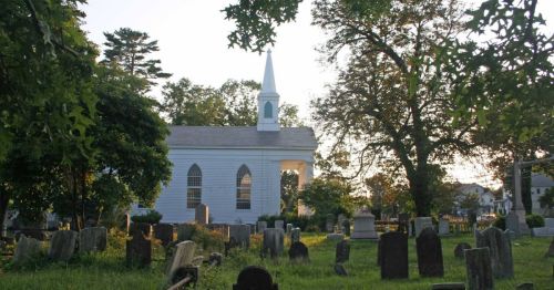 A serene graveyard with old tombstones, featuring a white church with a steeple in the background, surrounded by trees.