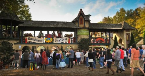 A lively crowd gathers at a Renaissance Festival, with performers on a stage and colorful banners in a festive setting.