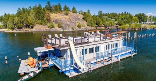 A floating house with a slide, surrounded by water and trees, featuring a rooftop deck and kayaks nearby.