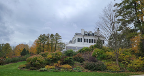A large white house on a hill surrounded by colorful autumn foliage and trees under a cloudy sky.
