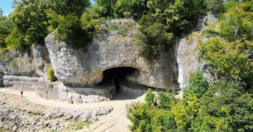 A rocky cliff with a large cave entrance, surrounded by lush greenery and a dirt path along the riverbank.