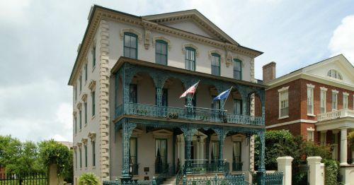Historic building with ornate ironwork balconies, featuring flags and lush greenery in the foreground.