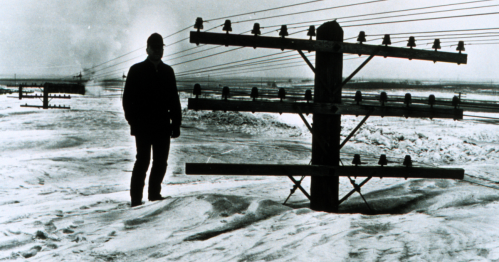A silhouette of a person stands beside a telephone pole in a snowy landscape, with power lines in the background.