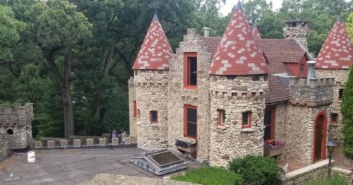 A stone castle with red-tiled roofs surrounded by trees, featuring turrets and a scenic outdoor area.