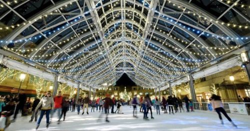 A bustling ice skating rink under a beautifully lit, arched structure adorned with twinkling lights.