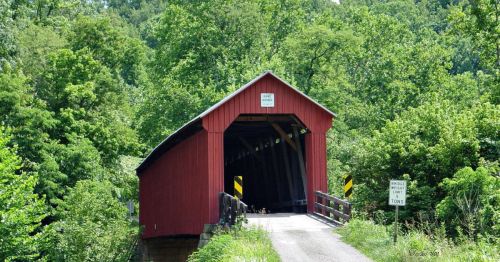 A red covered bridge surrounded by lush green trees, leading to a gravel road with caution signs.