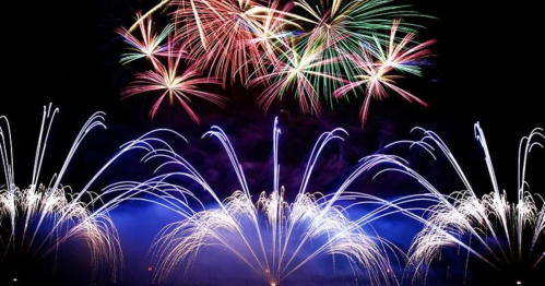 Colorful fireworks burst in the night sky, creating vibrant patterns above a shimmering display of lights.