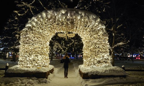 Woman walking under an arch of Christmas lights in a snowy park.