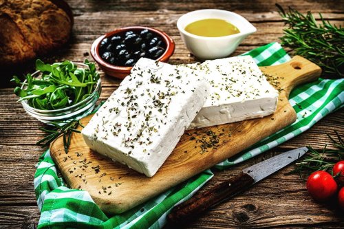 Blocks of feta cheese with herbs on a wooden board, surrounded by olives, olive oil, greens, and tomatoes.