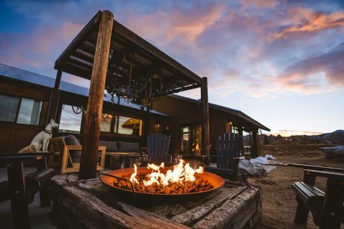 A cozy outdoor fire pit surrounded by wooden chairs, with a modern cabin and colorful sunset in the background.