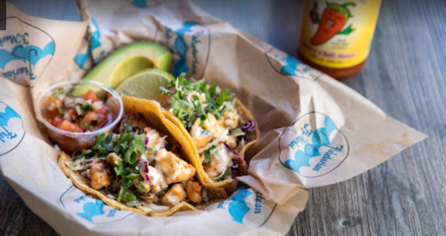 Two tacos filled with fish, topped with cabbage and salsa, served with lime and a bottle of hot sauce on the side.