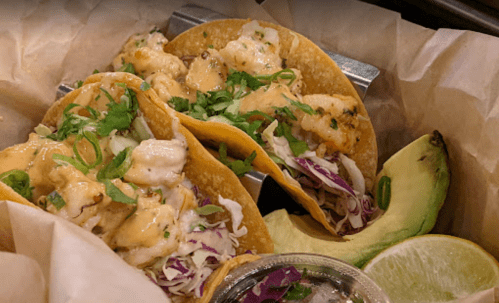 Three shrimp tacos topped with cabbage, cilantro, and sauce, served with avocado and lime on the side.